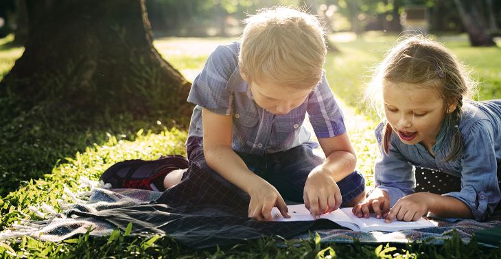 Kids sitting on a towel on the grass reading a notebook